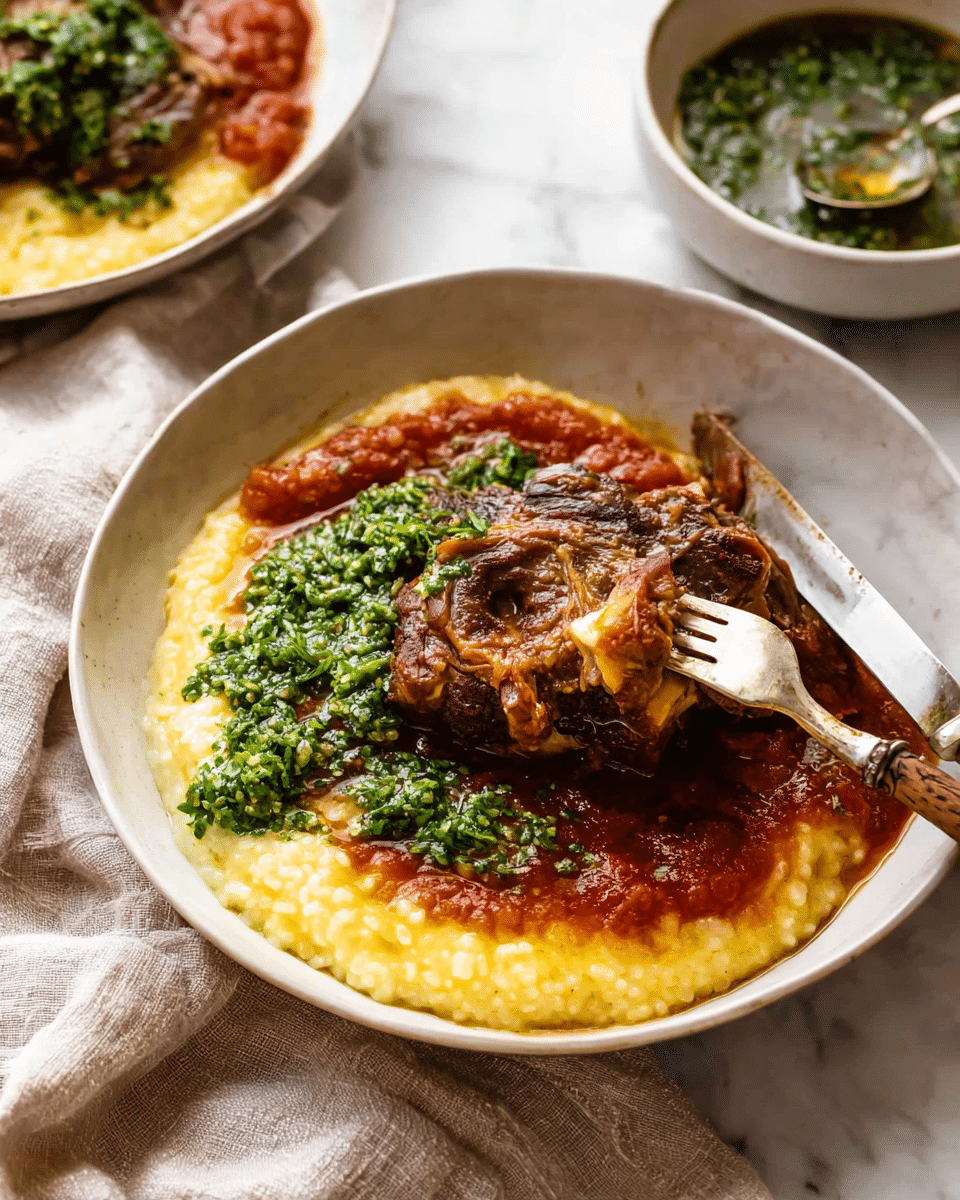 A close-up of a shallow white bowl with three distinct layers: the bottom layer is creamy and yellow risotto with a soft, thick texture visible; on top sits a large piece of brown braised meat with a round bone in the center showing marrow, covered in a rich red tomato sauce with a slightly chunky texture; the dish is finished with a dollop of bright green chopped herb garnish placed centrally on the meat, adding color contrast. A silver fork is stuck into the meat from the left side and a silver knife with a light wooden handle rests on the right edge of the bowl. Behind, a white bowl filled with finely chopped green herbs, with a spoon inside, is set on a soft beige cloth on a white marbled surface. photo taken with an iphone --ar 4:5 --v 7