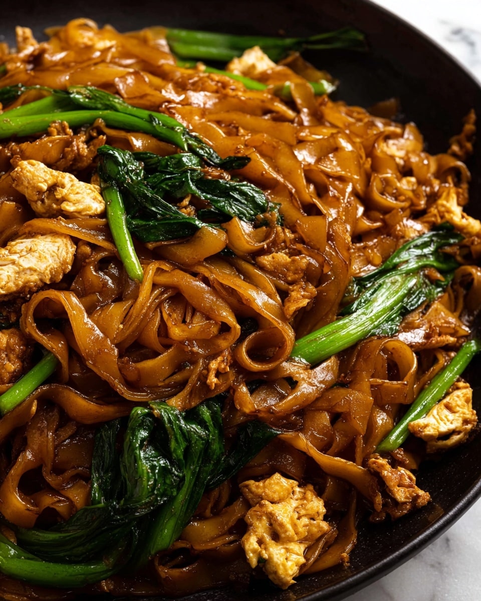 A close-up view of a dish featuring wide, flat noodles that are rich brown and glossy from sauce, mixed with small pieces of light brown cooked chicken and bright green leafy vegetables. The noodles are layered unevenly with some folded and curled, creating a textured look. The green leaves are scattered on top and around the noodles, adding contrast with their fresh, shiny surface. The dish is served in a white bowl, placed on a white marbled surface that adds subtle texture to the background. photo taken with an iphone --ar 4:5 --v 7