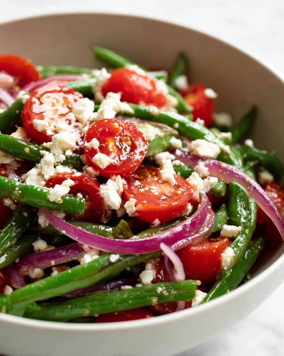 The image shows a close-up of a fresh vegetable salad in a white bowl. The salad has crisp green beans as the main layer, cut into smaller pieces and spread evenly. Mixed in are bright red cherry tomatoes cut in halves, adding a glossy texture and vibrant look. Thin slices of light purple onion rings are scattered throughout, adding a delicate layer. On top, there are small white crumbles of soft cheese sprinkled all over, creating a nice contrast with the green and red colors. The salad looks fresh and lightly dressed, with a subtle shine indicating a thin coating of dressing. The background is a white marbled texture. Photo taken with an iphone --ar 4:5 --v 7