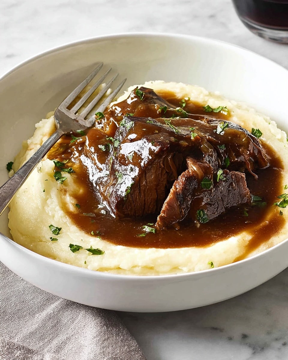 A white bowl contains a dish with two main layers: the bottom layer is creamy, pale mashed potatoes spread evenly in the bowl, and the top layer is a thick piece of dark brown meat covered in rich, glossy brown gravy. The meat is partially cut to reveal tender, pinkish fibers inside. Small green herb leaves are sprinkled over the gravy and mashed potatoes. A fork rests on the left side of the bowl, partially touching the mashed potatoes, and a knife is partly visible on the right side, cutting into the meat. The scene is set on a white marbled surface with a soft white cloth and a blurred dark glass in the background. Photo taken with an iphone --ar 4:5 --v 7