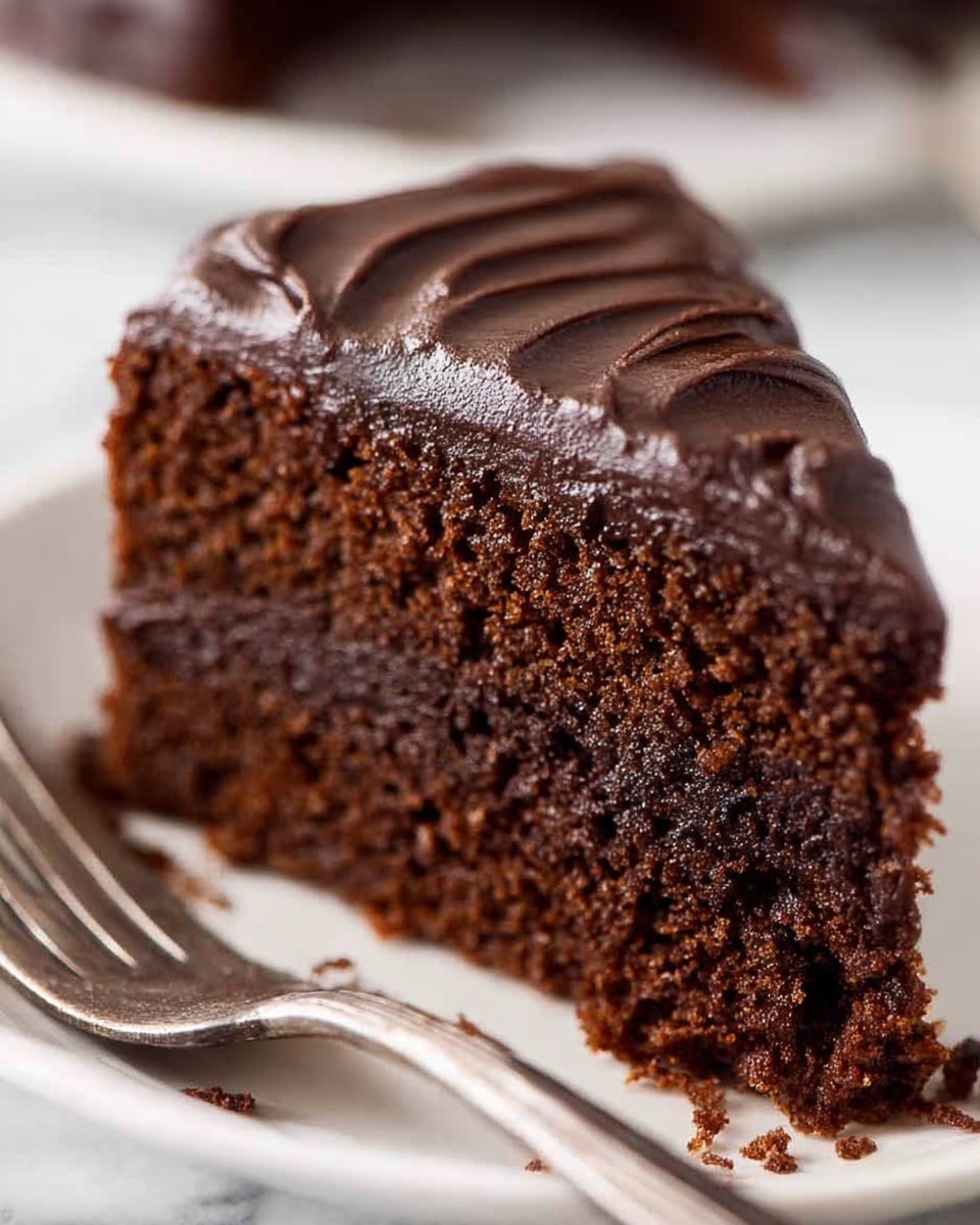 A close-up view of a single piece of chocolate cake with one thick layer of moist dark brown cake topped with a thick layer of dark, shiny, and smooth chocolate frosting, which has a slightly swirled texture. The cake is placed on a white plate on a white marbled surface. There are small crumbs scattered near the cake, and part of a fork is visible blurred in the background. photo taken with an iphone --ar 4:5 --v 7